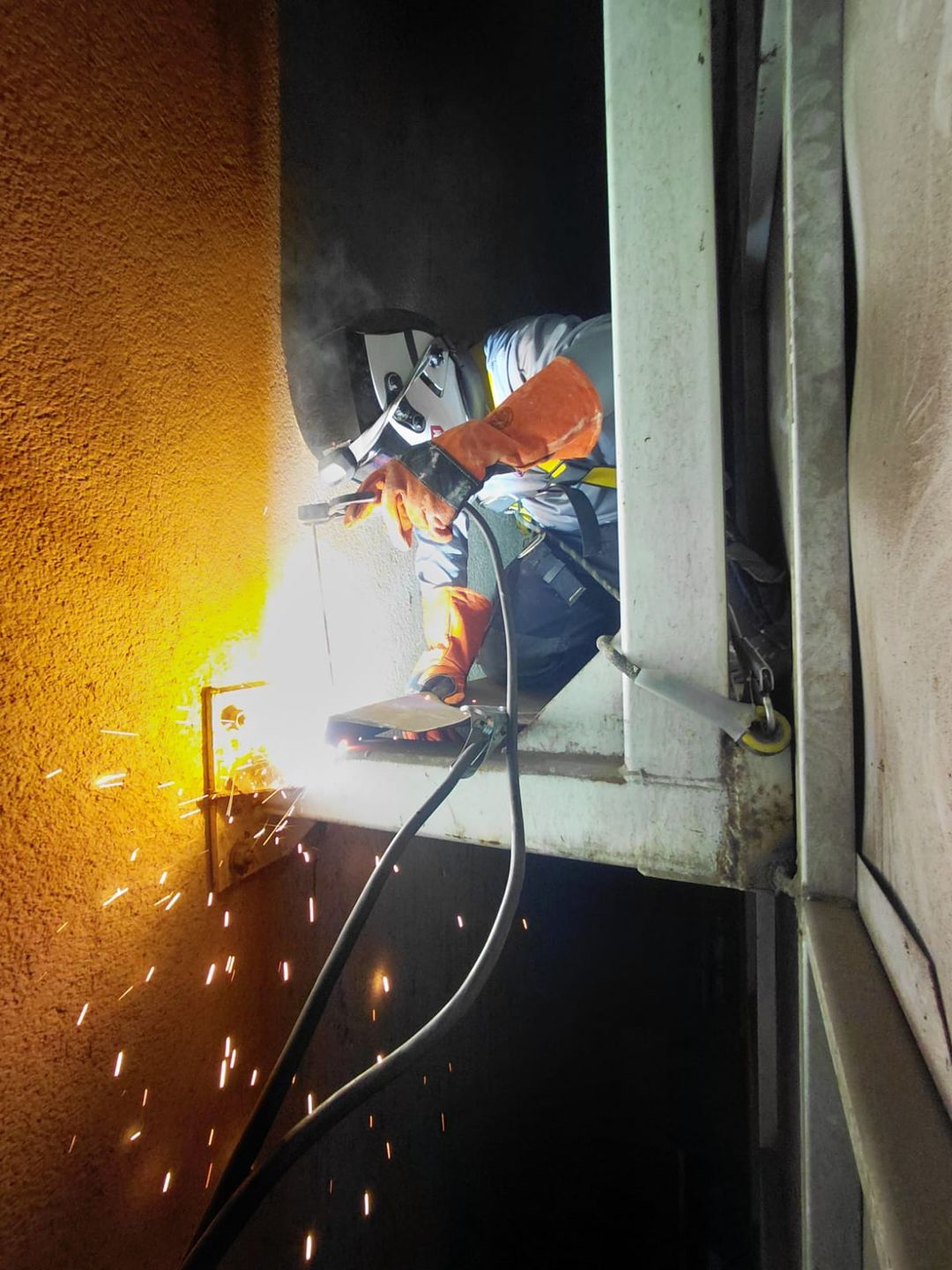 Soldador con arnés y equipo de protección soldando una estructura metálica en altura, con chispas cayendo